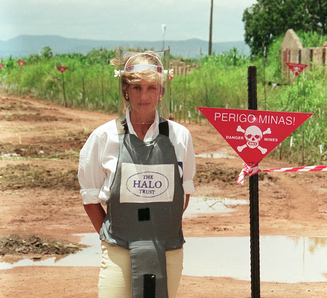 Image of a blonde woman in a bulletproof vest and clear face shield standing in a middle of a dirt field next to a red sign with a skull of bones