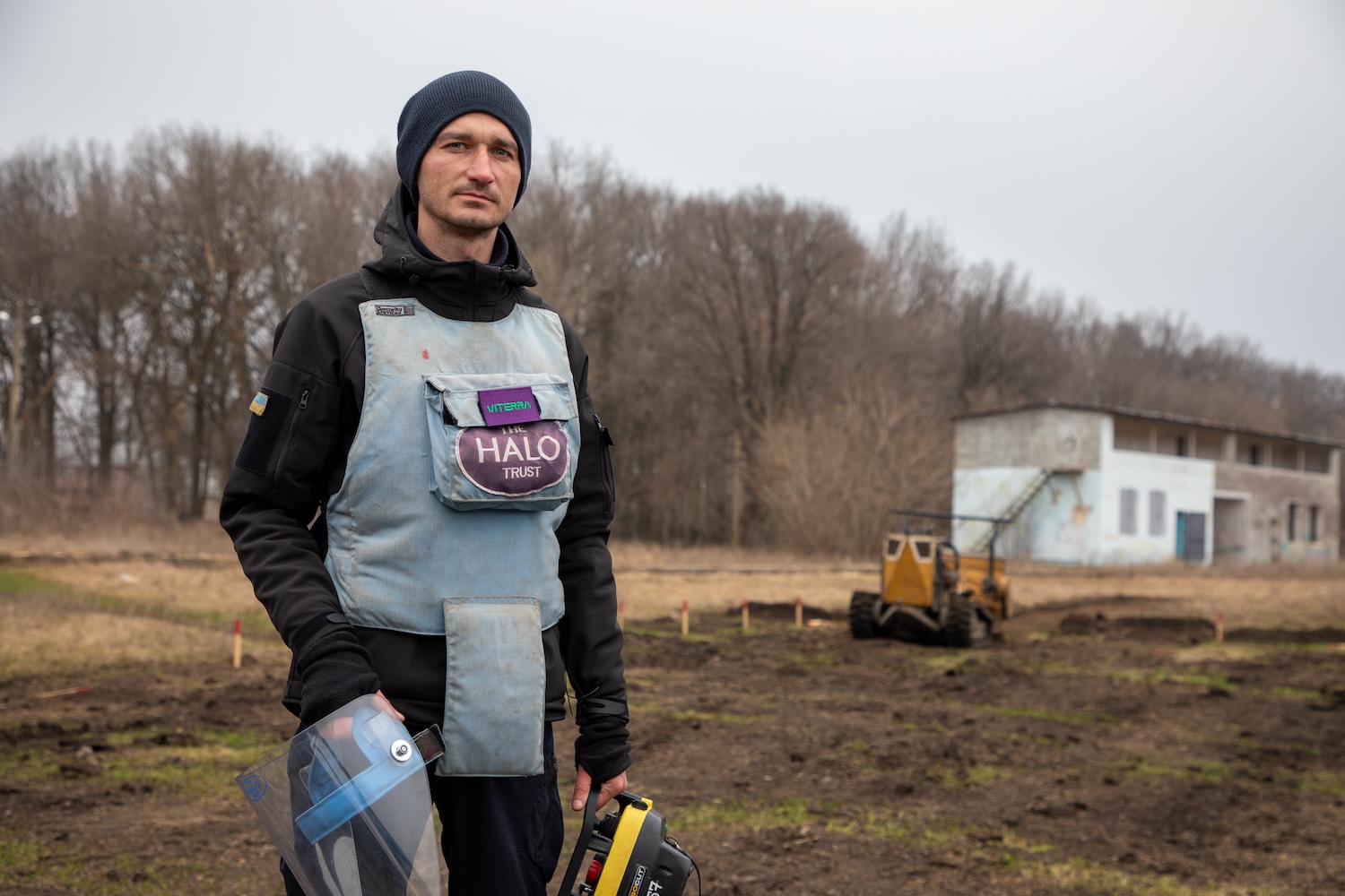 Image of a man wearing all black with a light blue protective vest that says "Viterra" above "The HALO Trust." He stands in a field where the grass is dead and the trees are bare.