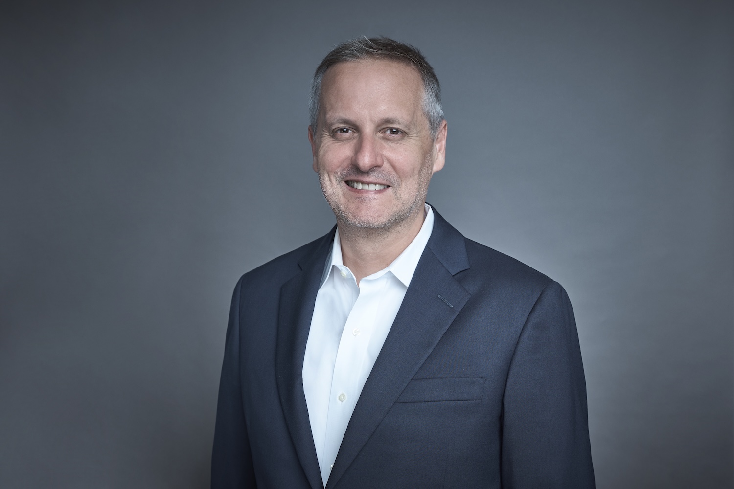 Portrait of a middle-aged man in a dark suit jacket and white shirt, smiling against a gray background.