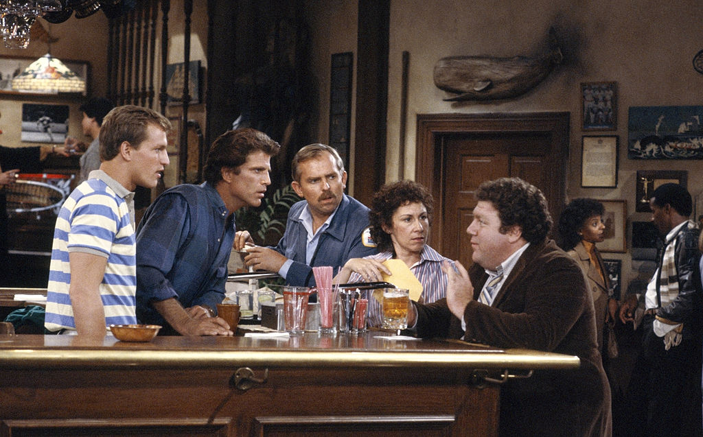A scene from a television sitcom showing several characters gathered at a bar, leaning in toward one another as they talk, with other patrons and bar décor in the background.