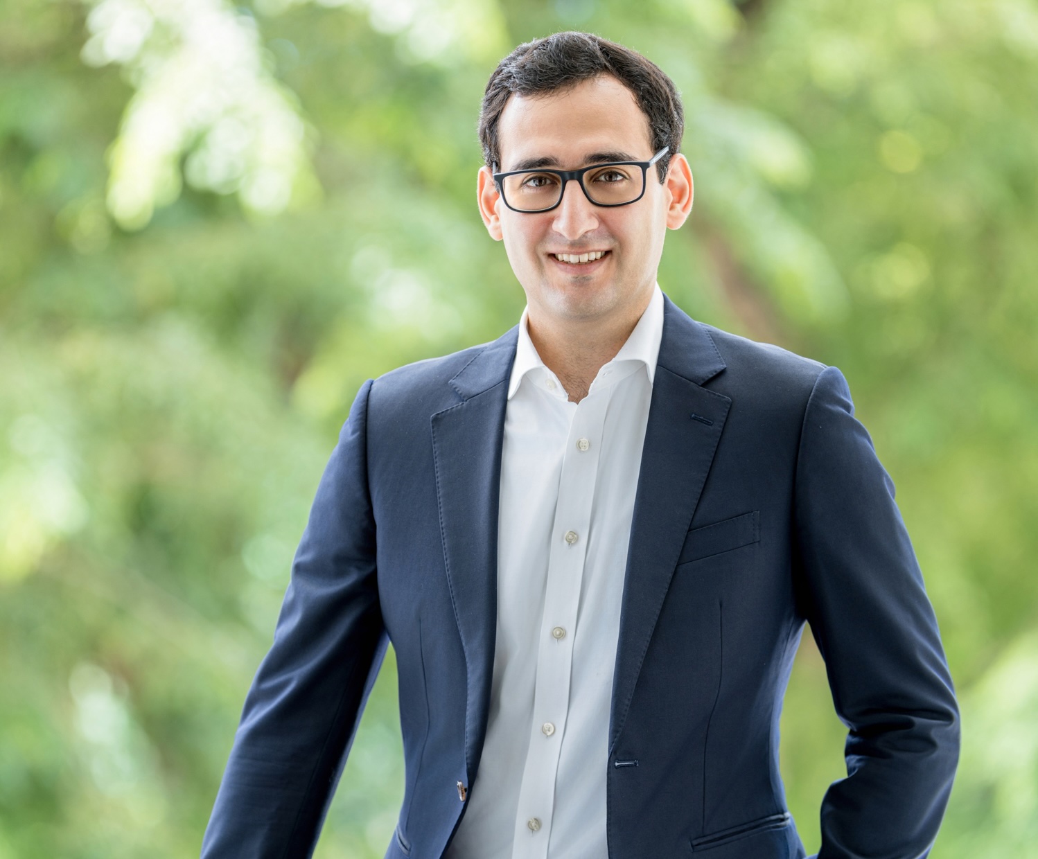 a man in a navy suit jacket and white button down with black-rim glasses standing in front of green bushes or trees.