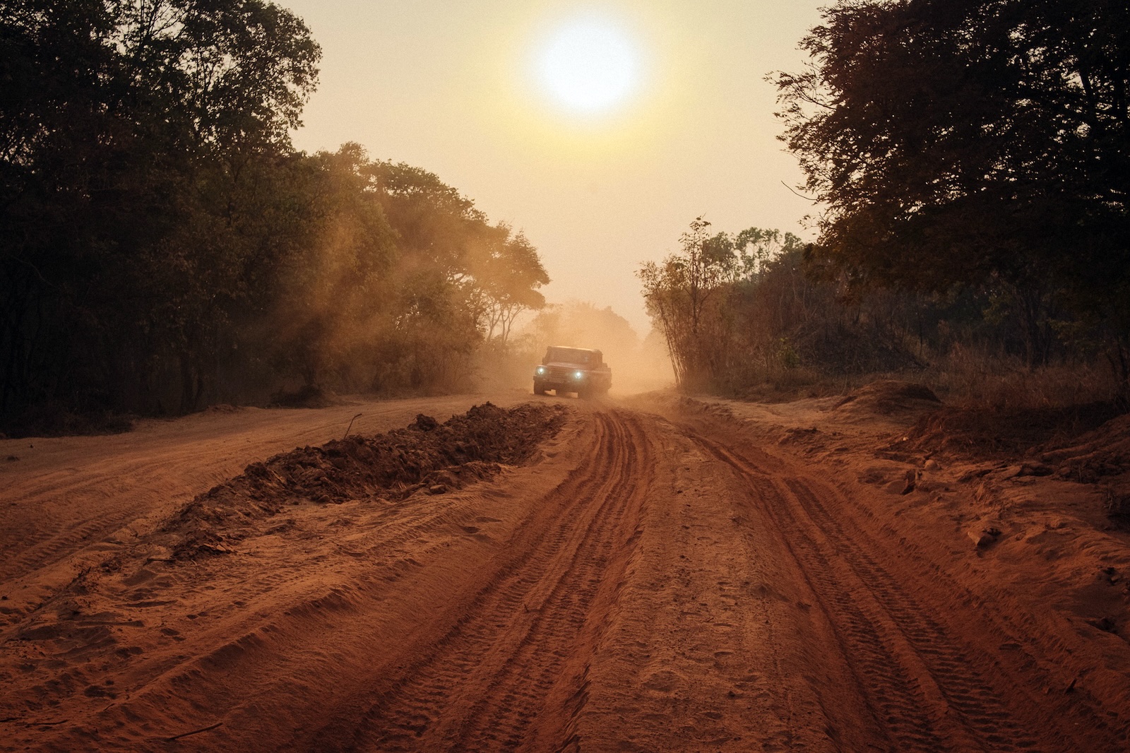 an image of an SUV driving down a dirt road lined with trees while the sun is descending behind it