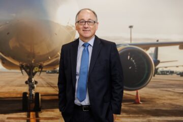 A man in a navy suit and royal blue tie wearing black rimmed glasses standing in front of a backdrop of an airplane