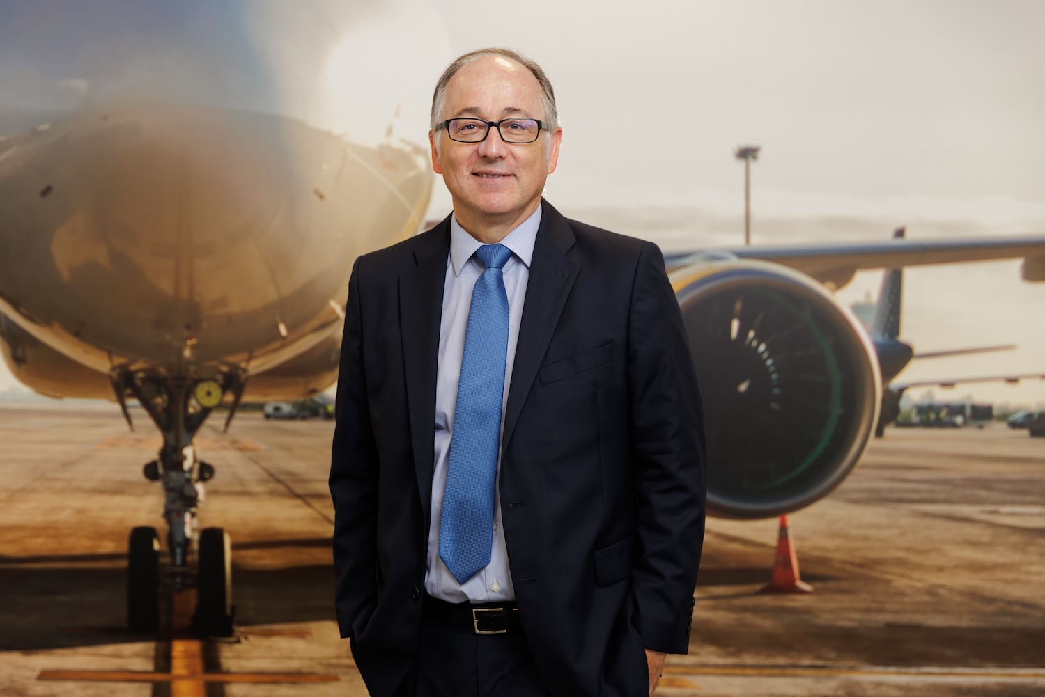 A man in a navy suit and royal blue tie wearing black rimmed glasses standing in front of a backdrop of an airplane