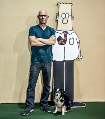 Scott Adams, a man wearing jeans and a teal t-shirt and black-rimmed glasses, standing next to a lifesize cutout of Dilbert the cartoon. An australian shepherd dog stands in front of them.