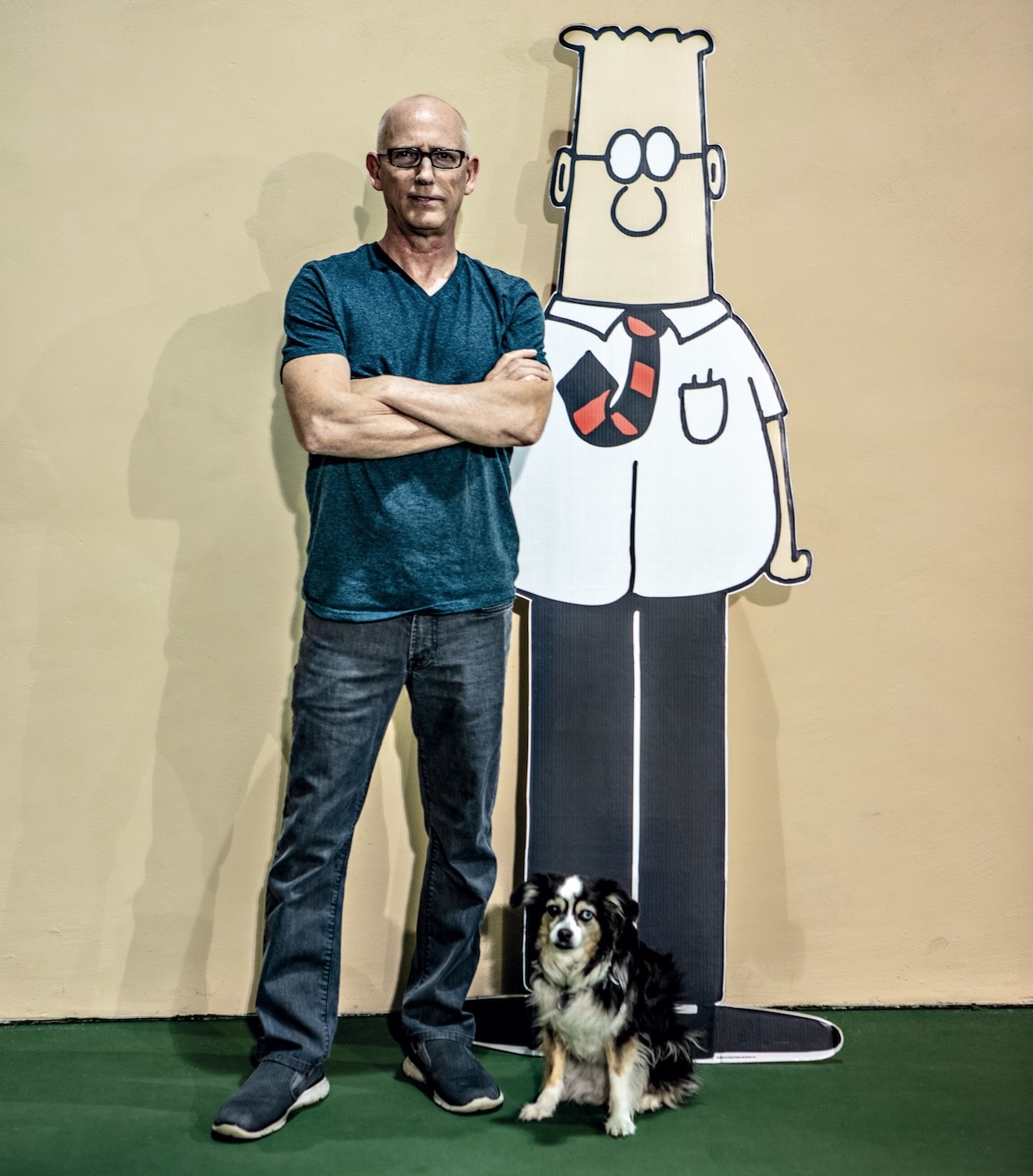 Scott Adams, a man wearing jeans and a teal t-shirt and black-rimmed glasses, standing next to a lifesize cutout of Dilbert the cartoon. An australian shepherd dog stands in front of them.