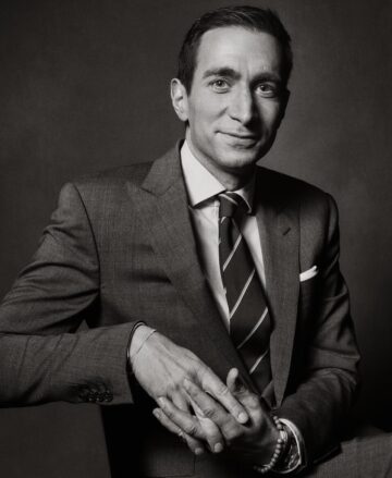 A black and white image of a man wearing a gray suit and white shirt with a striped tie. He's sitting on a chair with his hands clasped around the corner.