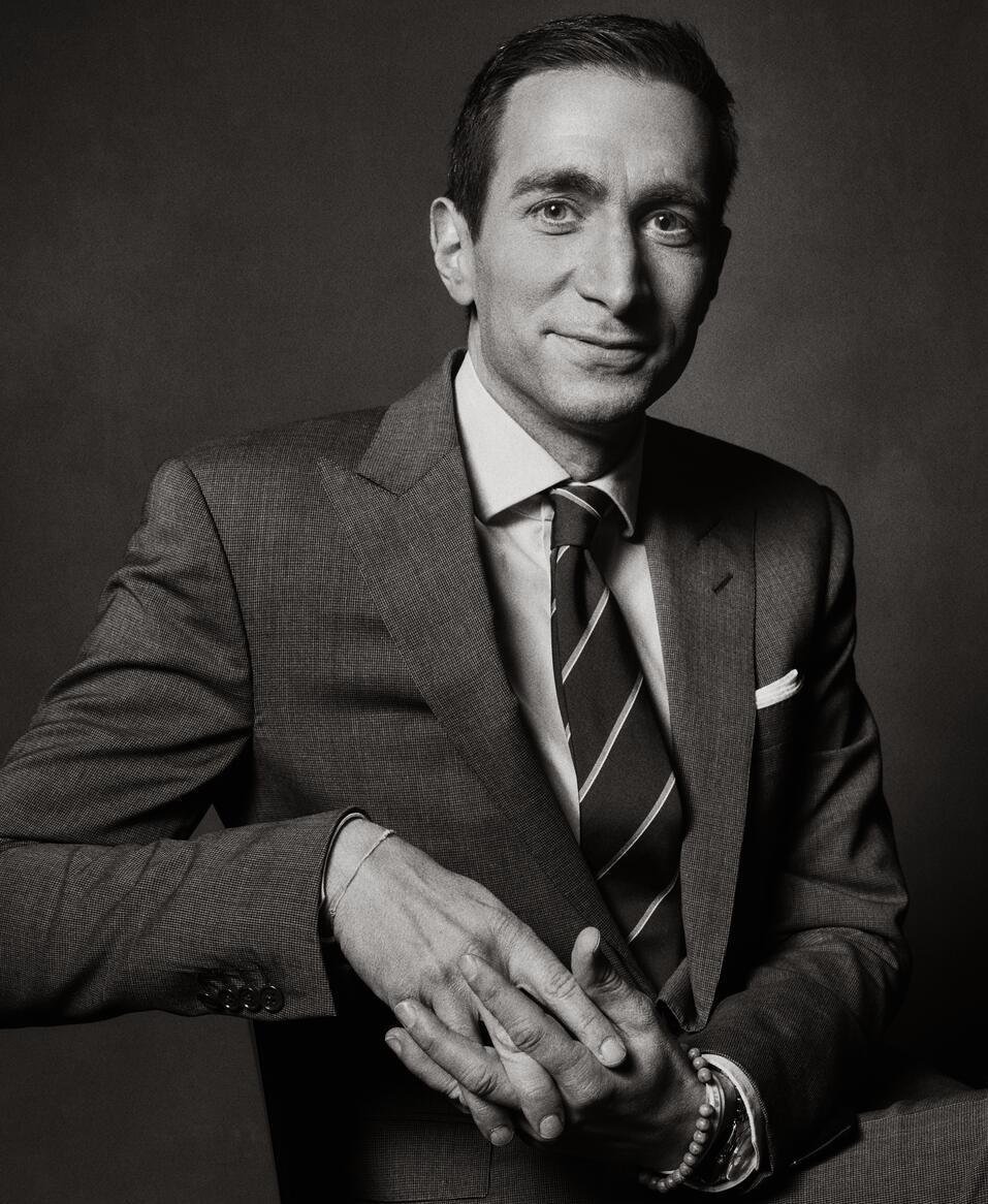 A black and white image of a man wearing a gray suit and white shirt with a striped tie. He's sitting on a chair with his hands clasped around the corner.