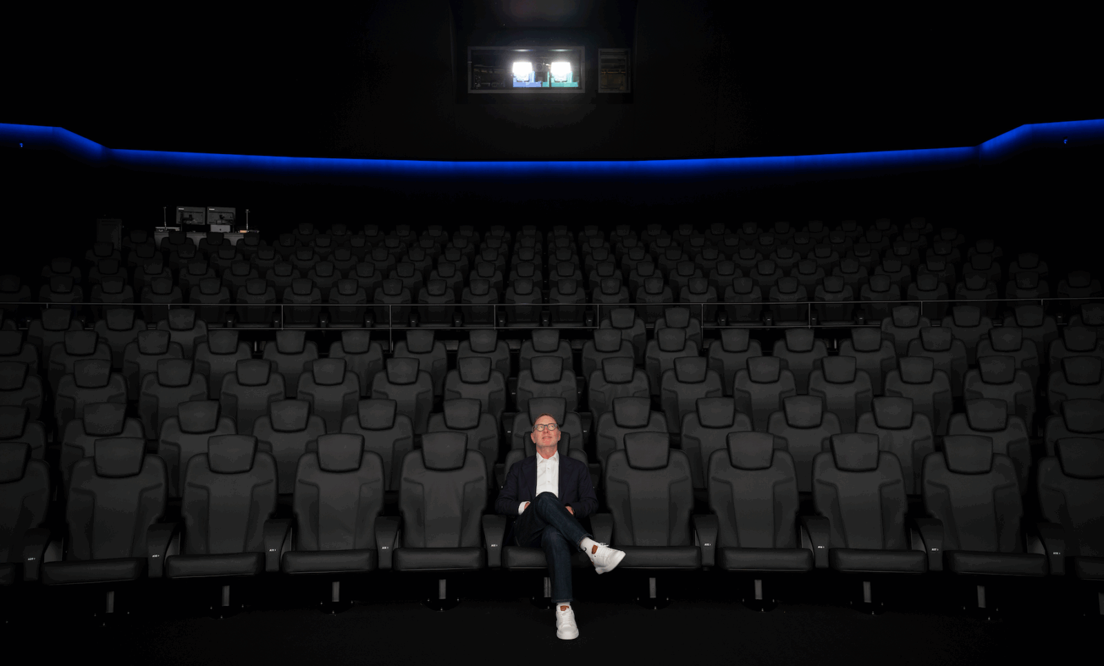 image of a man sitting alone in an empty theatre in the center front row looking up at the screen (behind the camera) in awe