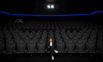 image of a man sitting alone in an empty theatre in the center front row looking up at the screen (behind the camera) in awe
