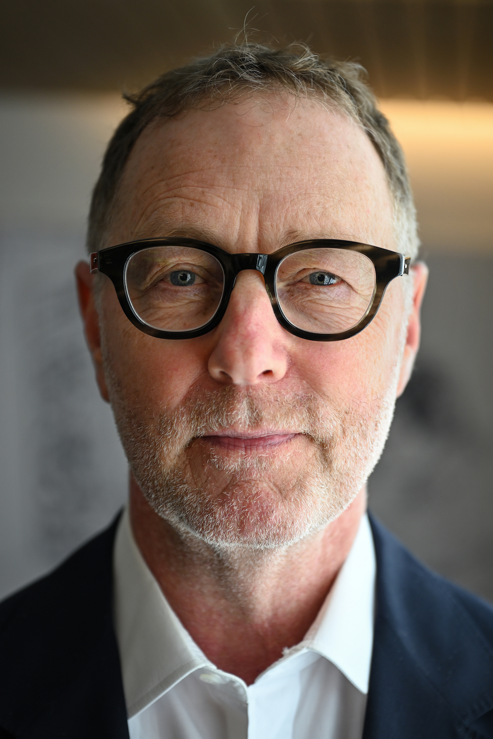 a close up photograph of a man wearing black rimmed glasses and a white button down and navy blazer looking straight into the camera