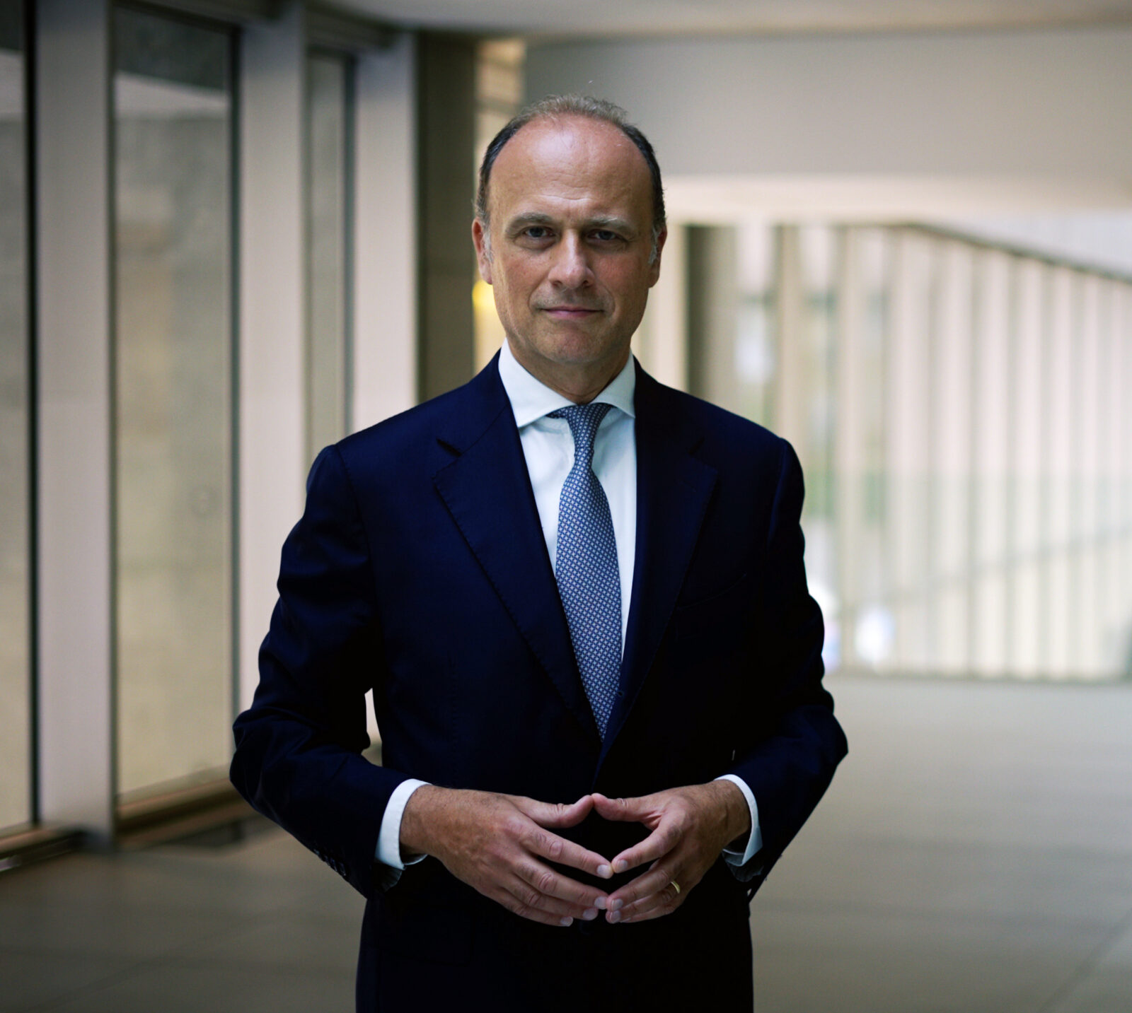 a photograph of a man in a dark blue suit with a blue tie standing in an administrative building lined with windows