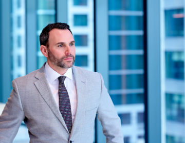 a man in a gray suit standing in front of glass windows with turquoise buildings in the background