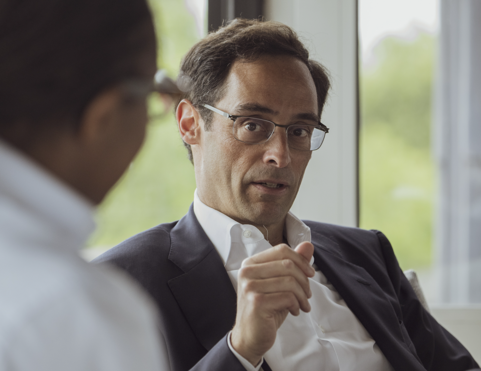 an image of a man in black rimmed glasses wearing a suit jacket and white button down, mid- conversation to someone whose back is to the camera and is slightly out of frame