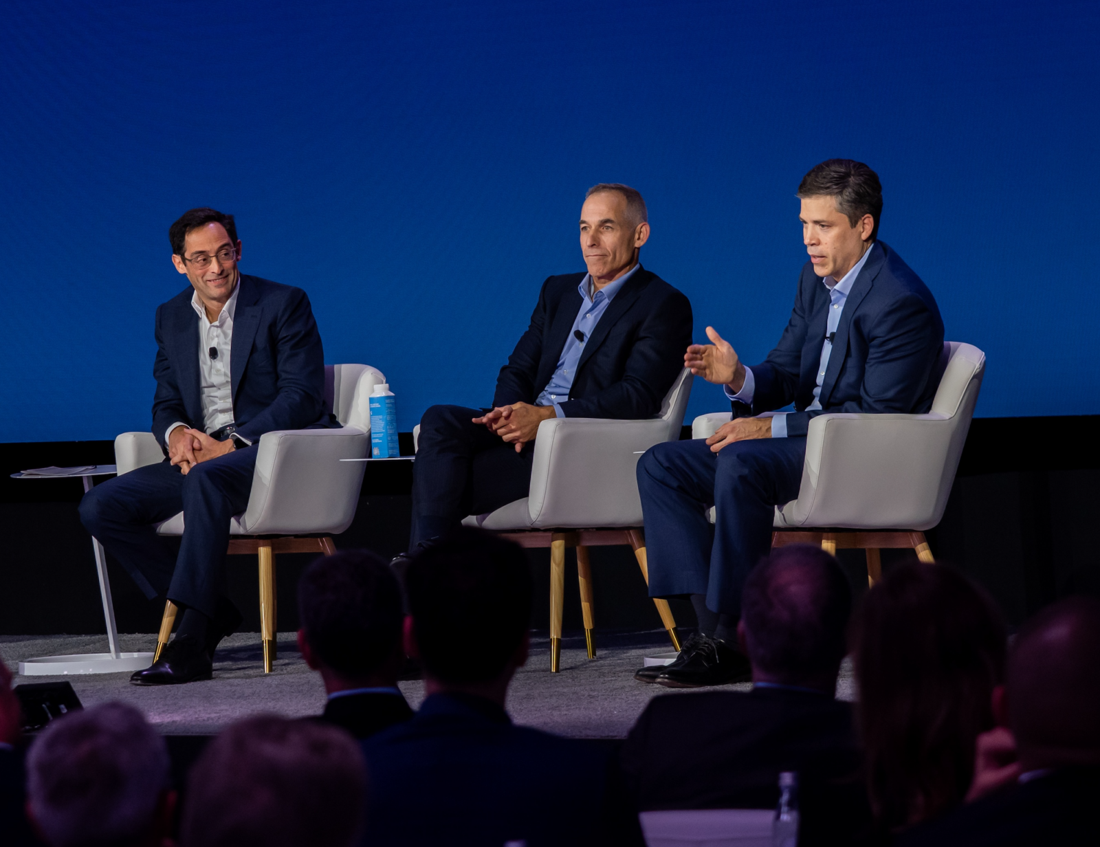an image of three men in suit jackets sitting in gray chairs on a stage with a blue screen in the background