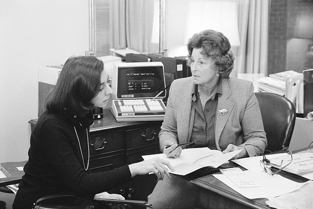 a black and white photograph of two women with a very old version of a computer behind them, discussing paperwork in front of them.