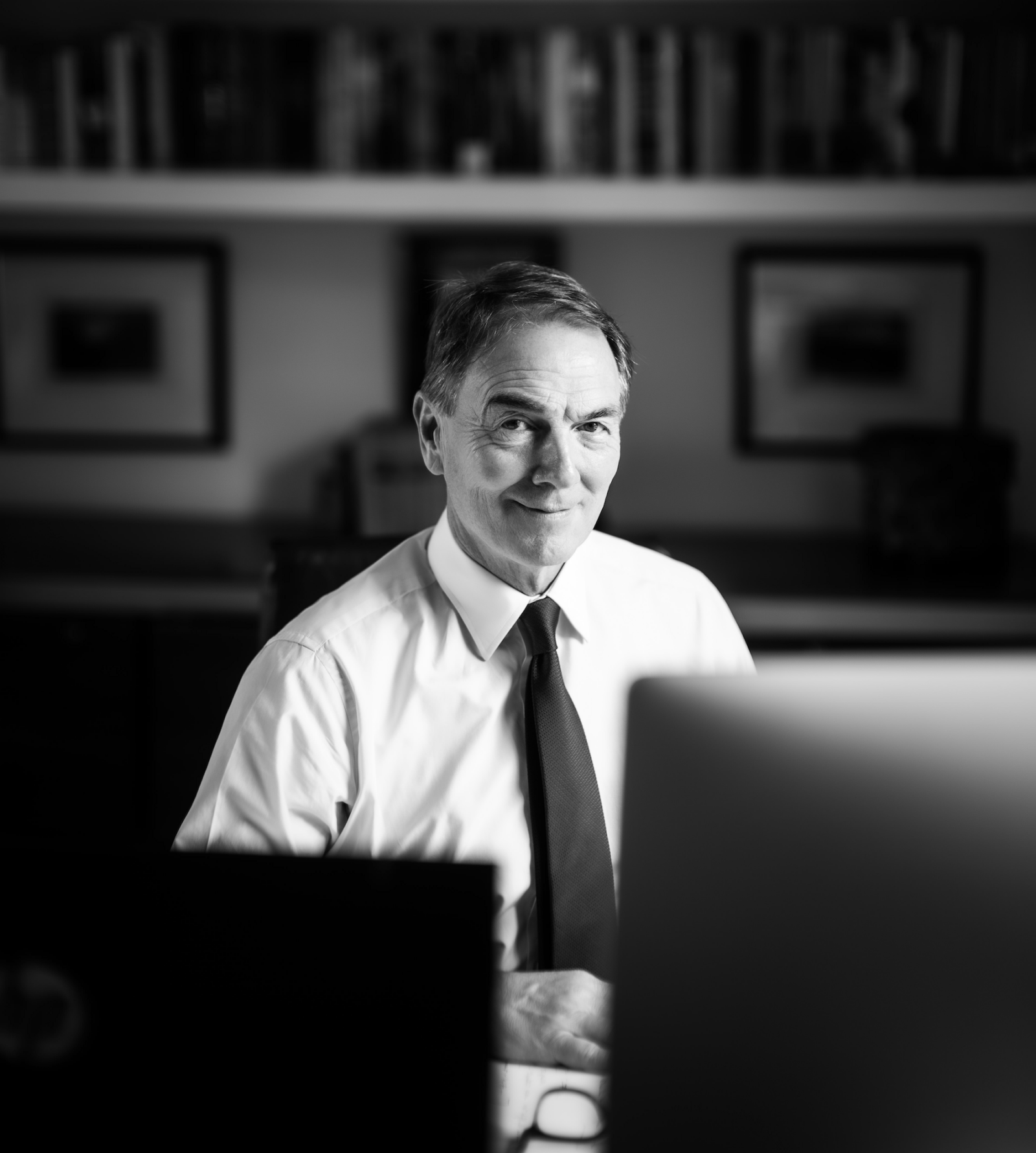 a black and white photo of a man in a shirt & tie sitting at a desk with two computer screens in the foreground