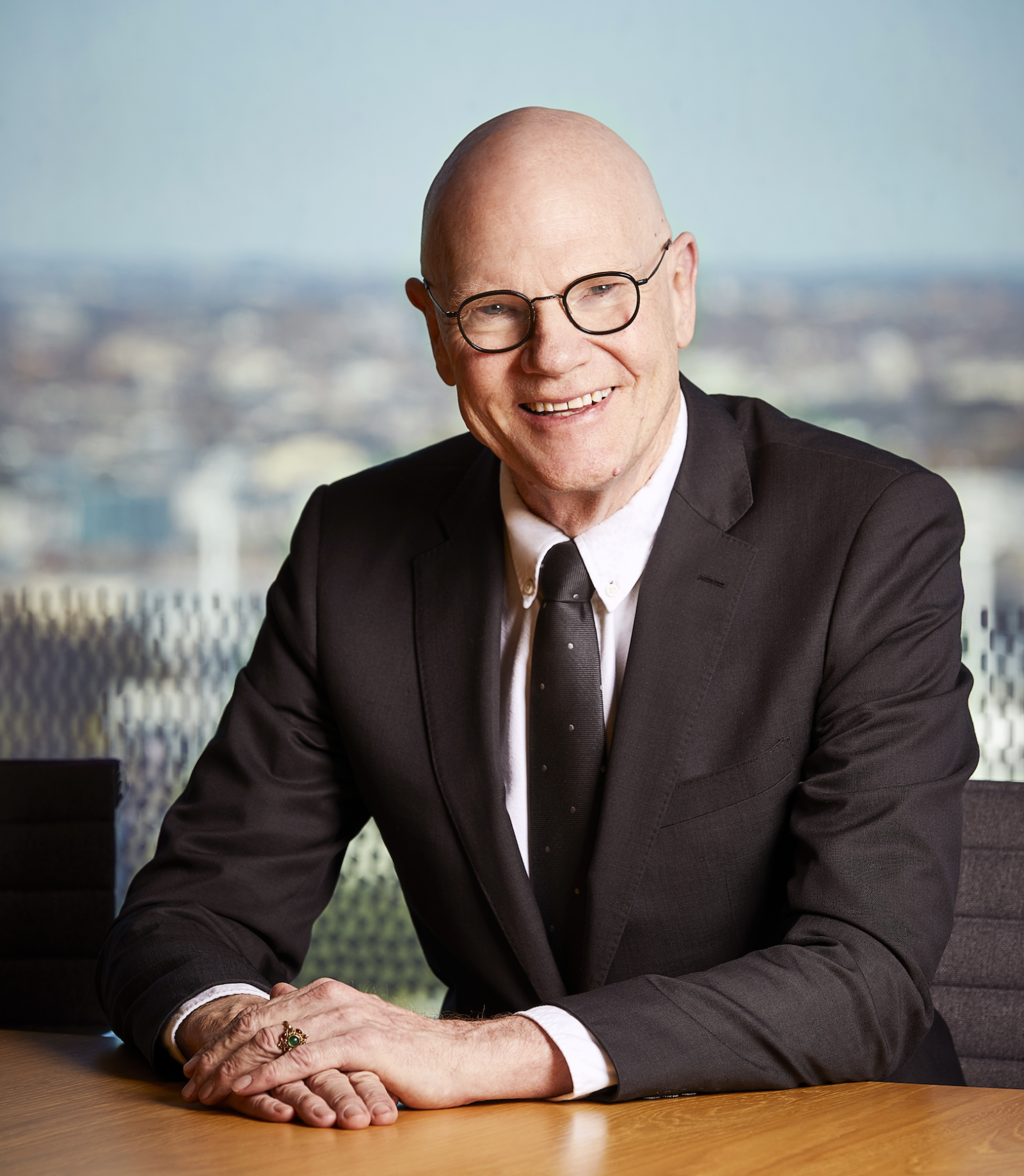 a photograph of a bald man with glasses wearing a suit sitting at a desk with a large window behind him