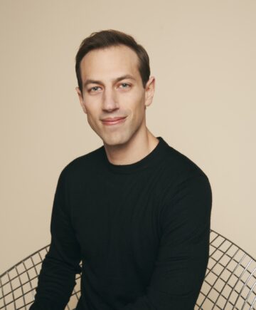 photograph taken of a man in a black sweater smiling at the camera siting in a wire chair with a tan backdrop behind him