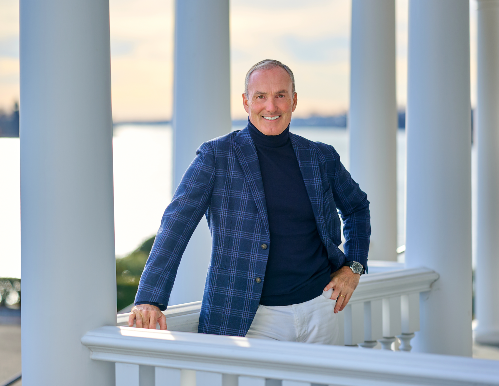 a photograph of a man in a black turtleneck, white pants and a blue plaid jacket standing on a deck with a body of water behind him