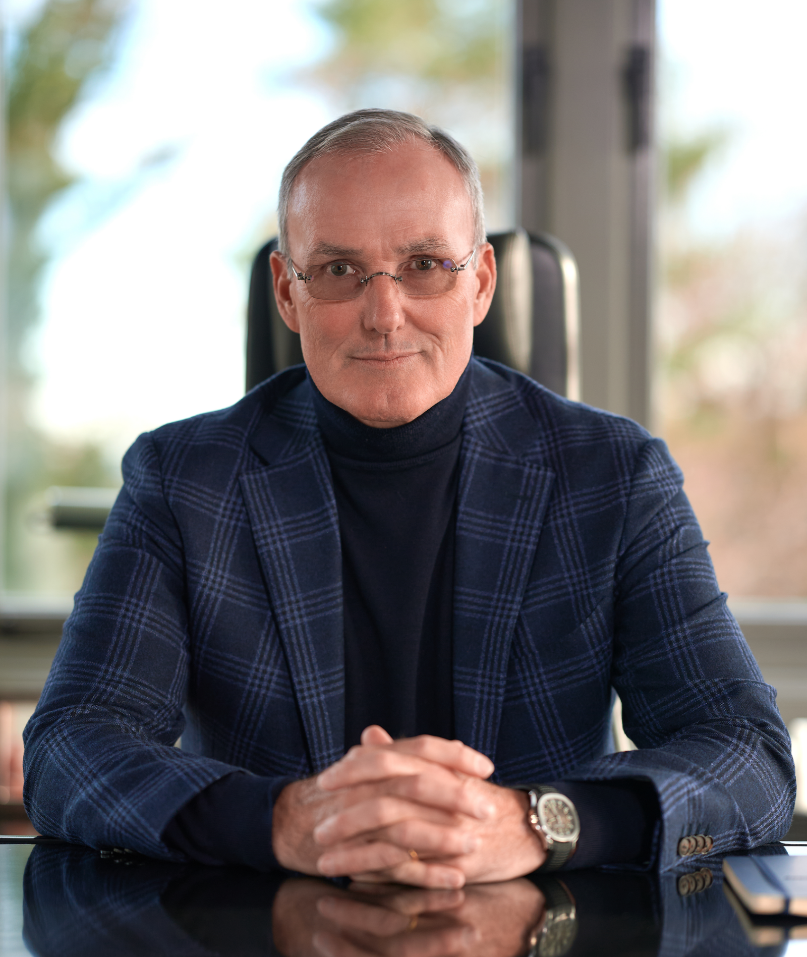 a man sitting at a desk in a navy plaid suit jacket and black turtleneck with his hands folded, he wears transition sunglasses