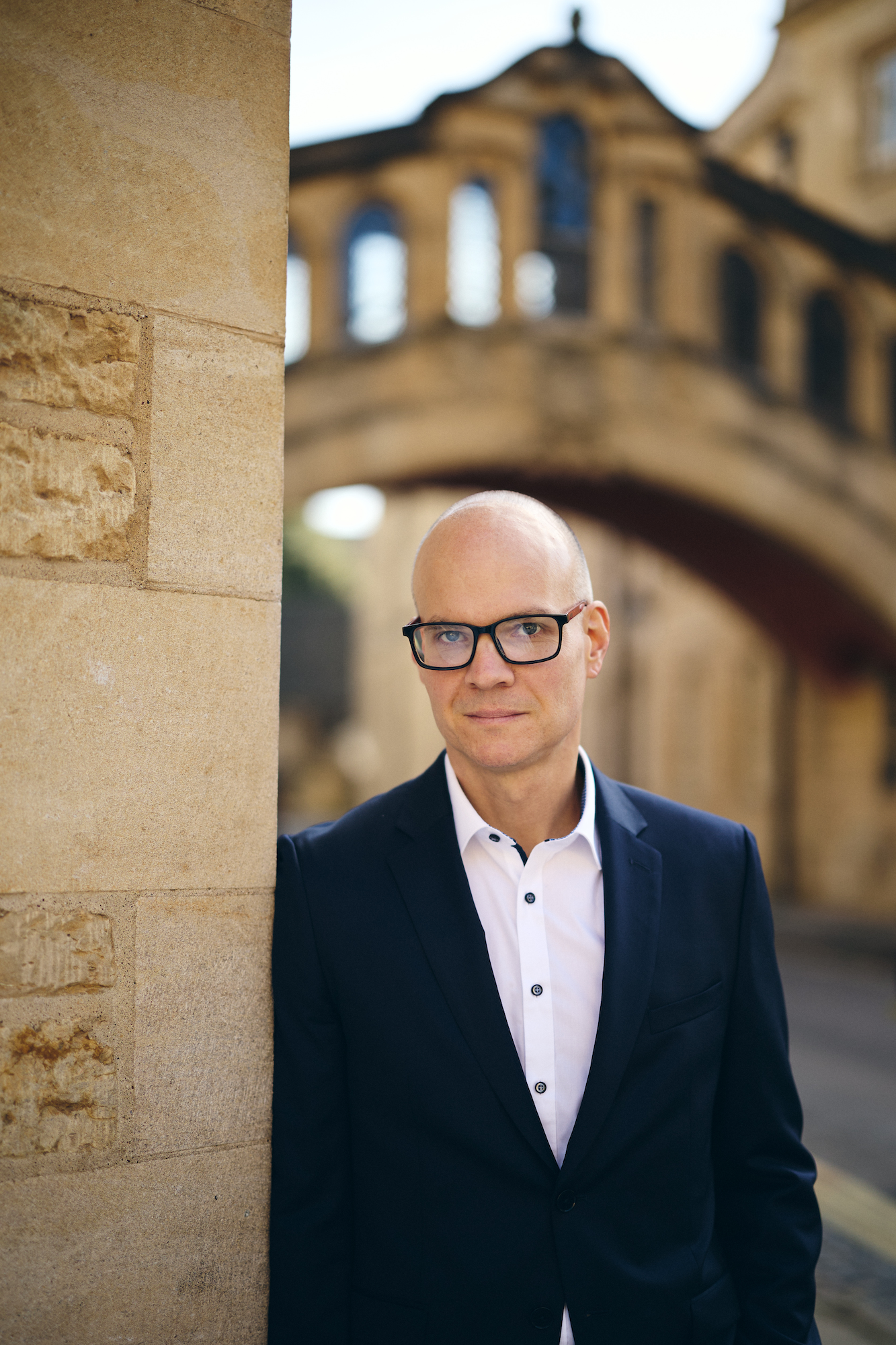 a man in black-rimmed glasses and a navy suit jacket standing beside a tan building with an archway behind him
