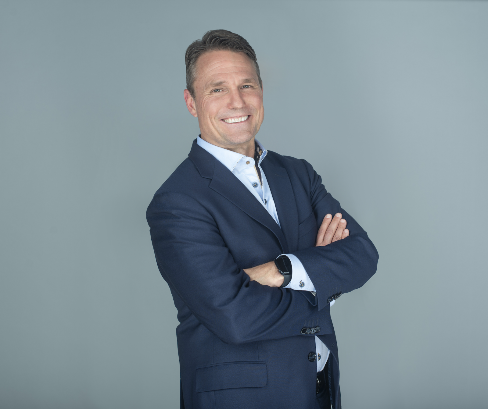 a photograph of a man in a navy suit with a big smile and his arms crossed across his chest standing in front of a gray backdrop.