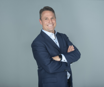 a photograph of a man in a navy suit with a big smile and his arms crossed across his chest standing in front of a gray backdrop.