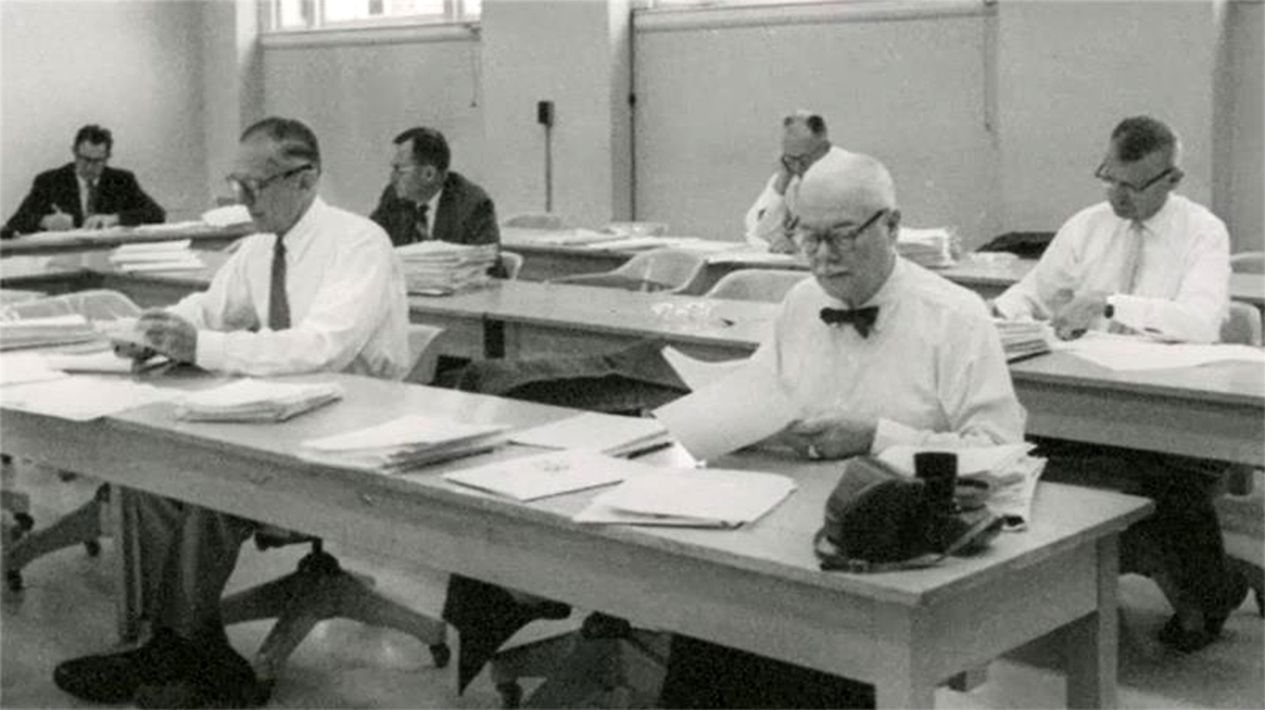 a black and white photo of men in white shirts & ties witting at desks with papers in front of them
