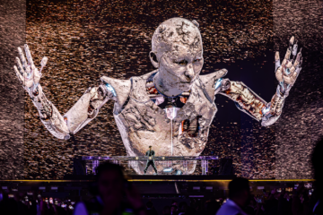 a photograph of a DJ performing on stage with a large robotic image on an LED screen in the background