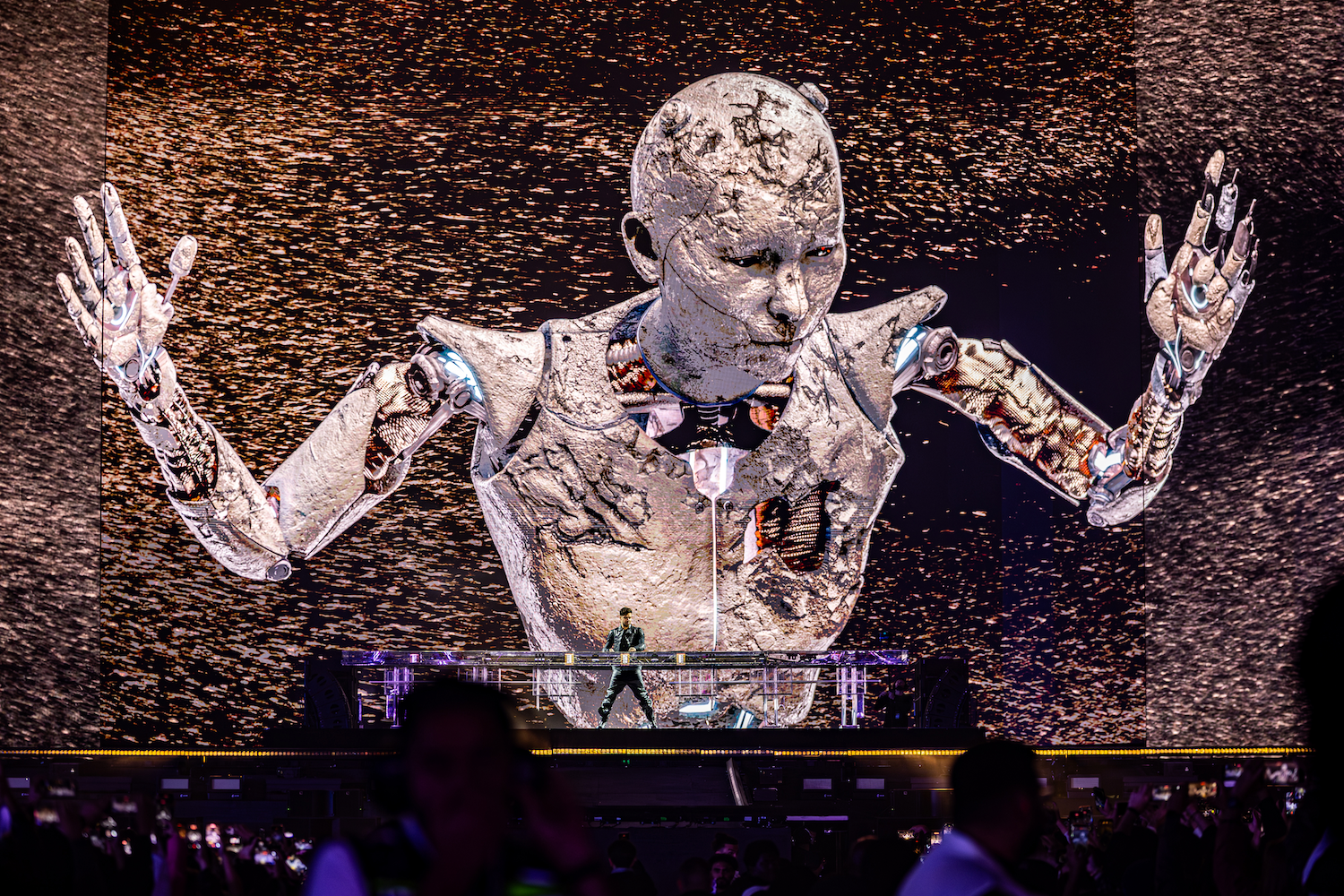 a photograph of a DJ performing on stage with a large robotic image on an LED screen in the background