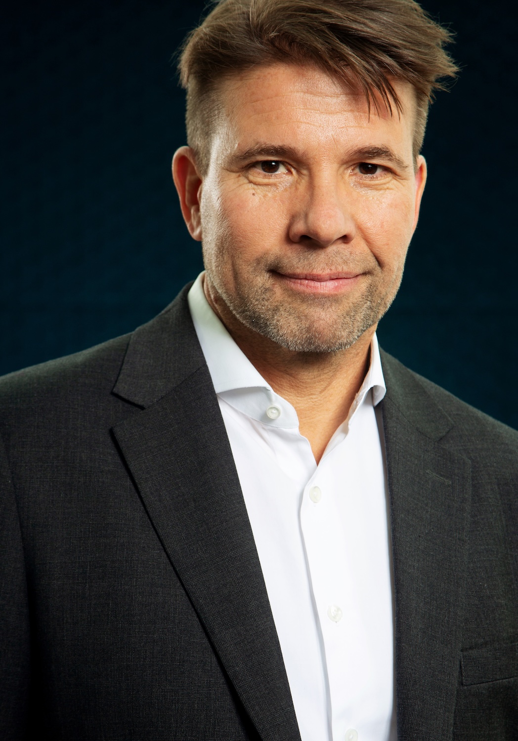 a photograph of a man in a gray suit jacket and white shirt smiling for the camera