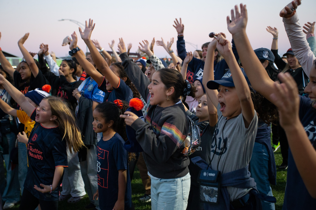 a photograph of a crowd of young giels wearing t-shirts and sweatshirt with the Bay Collective logo, all cheering with their hands raised high and mouths open.