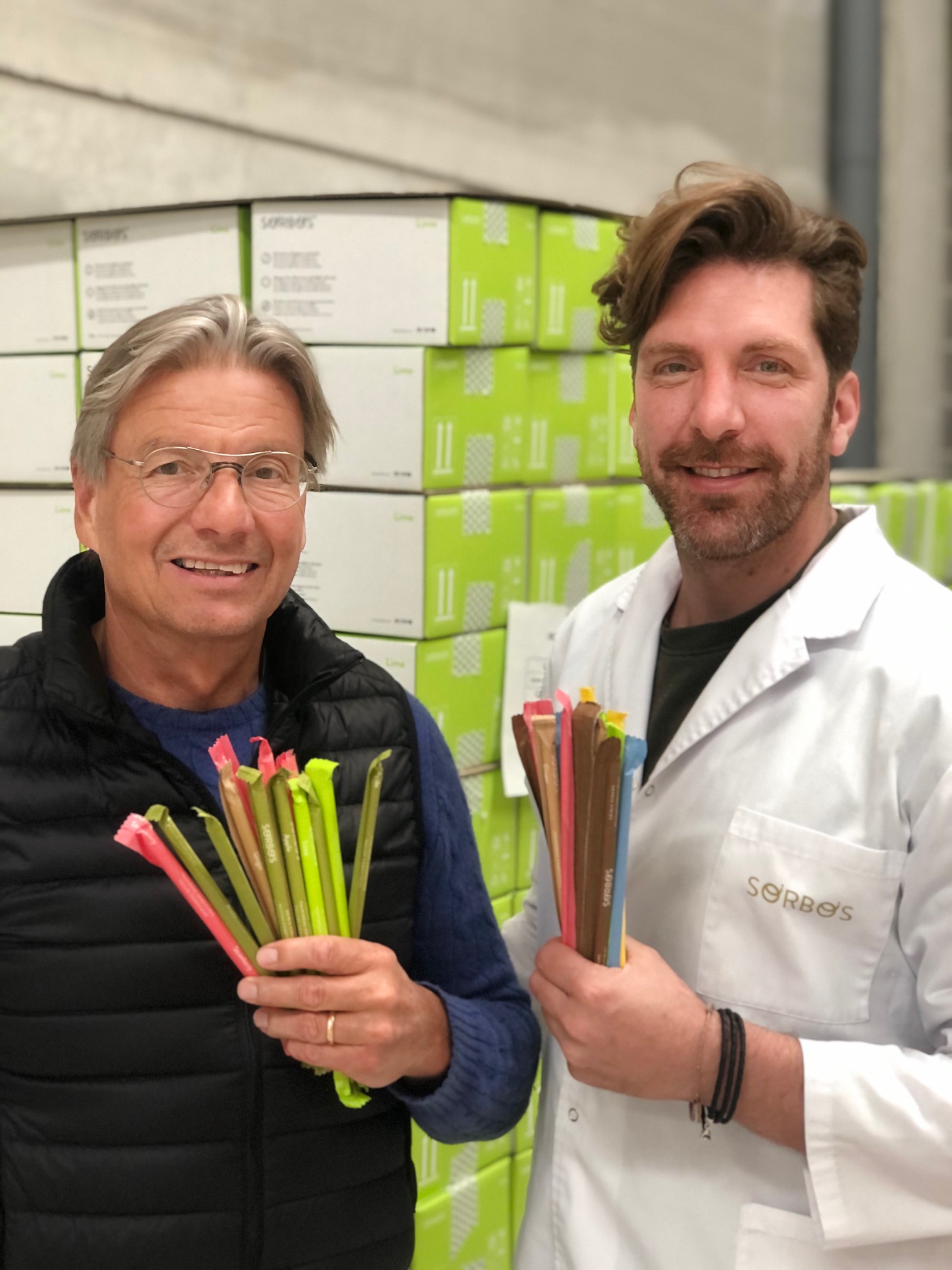 photograph of two men, one with gray hair in a black puffy vest, one younger and in a white lab coat, smiling for the camera holding handfuls of straws in their right hands.