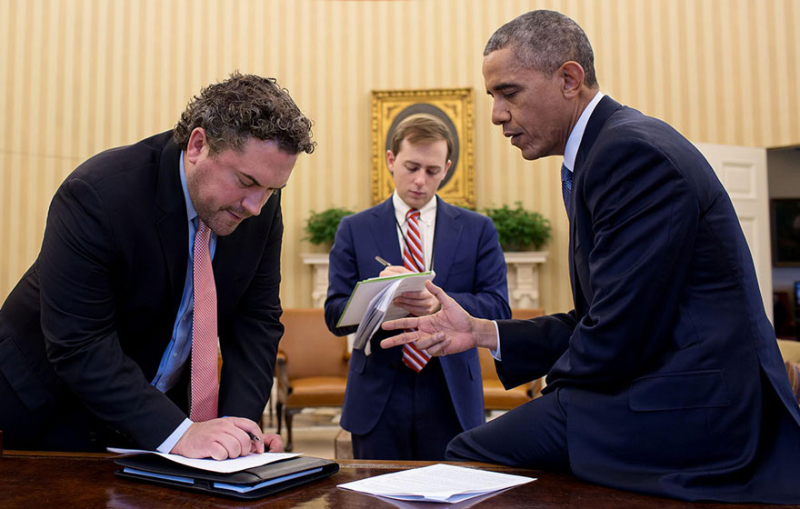 a picture of three men in the oval office bent over a piece of paper, one of the men is writing on it.