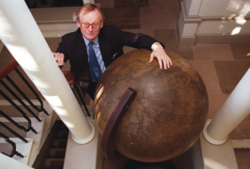 a photo of a man standing at the bottom of a staircase, taken from above, he has his hand on a giant wood globe.