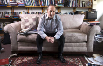 a photograph of a man sitting on a gray couch with a bookshelf behind it. he is leaning on his knees with his hands clasped.