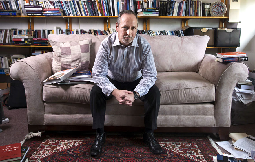 a photograph of a man sitting on a gray couch with a bookshelf behind it. he is leaning on his knees with his hands clasped.