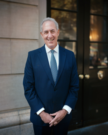 photograph of a man in a navy suit smiling outside a building