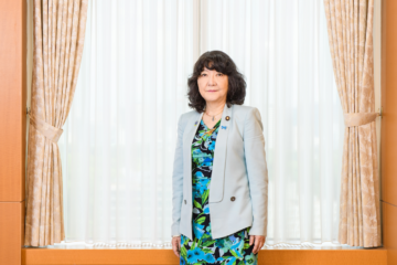 a Japanese woman in a suit jacket and floral dress standing in front of a curtained window