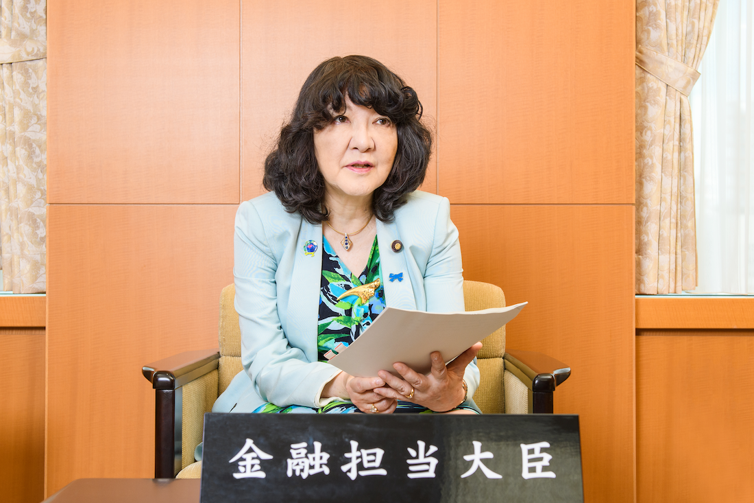 a Japanese woman in a suit jacket sitting in front of a podium reading from a booklet